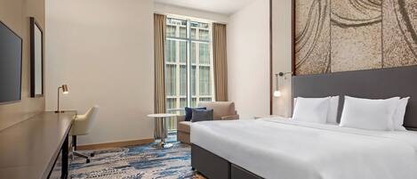 A hotel room with a large bed, a desk, a chair, a small table, and a window with curtains.