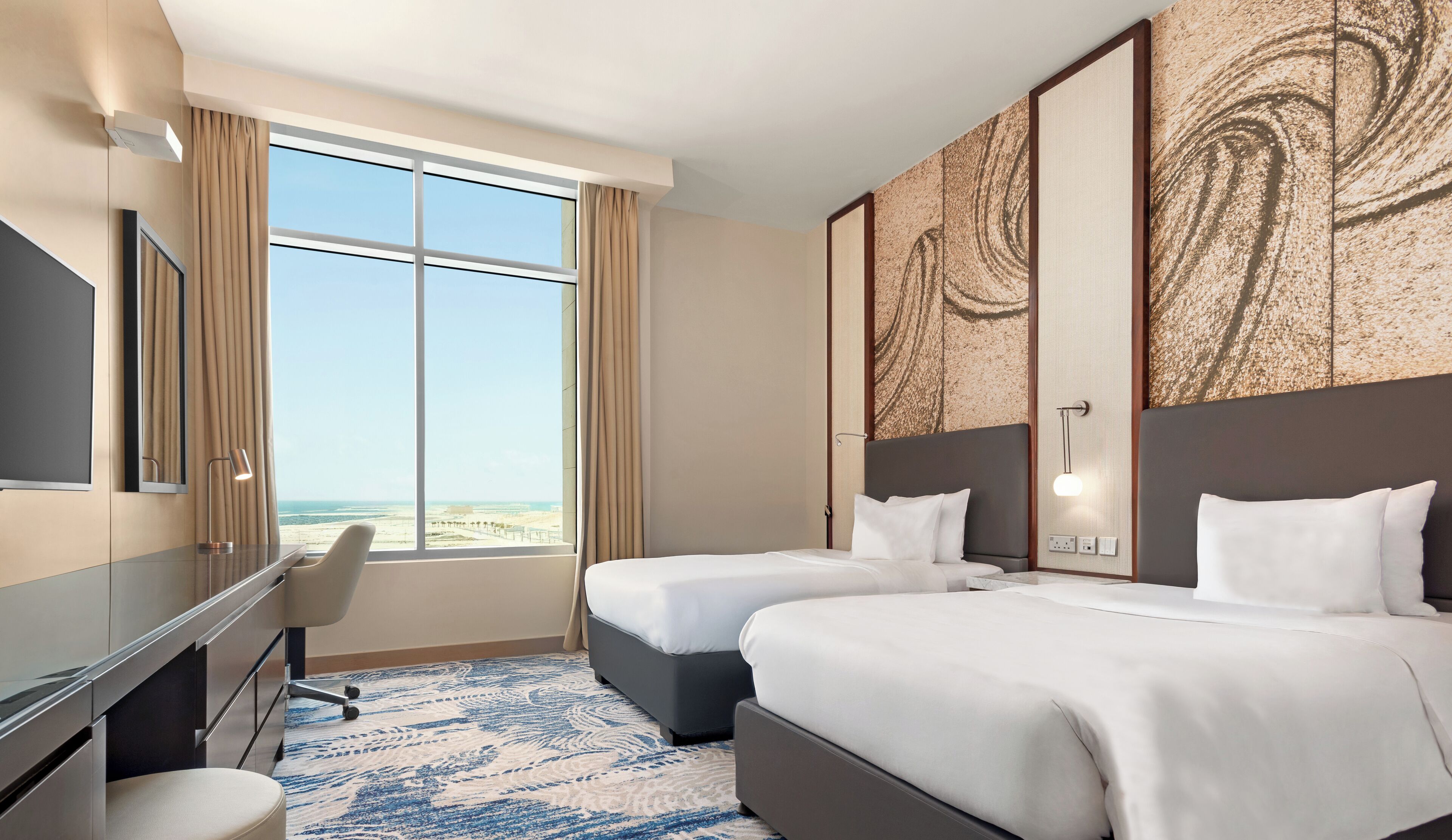 Habitación de hotel con dos camas, un escritorio, una silla y un amplio ventanal con vista a la playa.