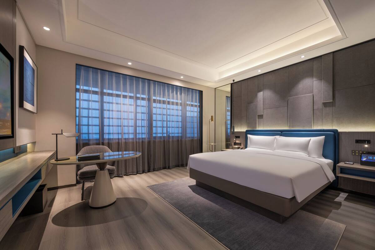 A modern hotel room with a large bed, a desk, a chair, and a large window with curtains.