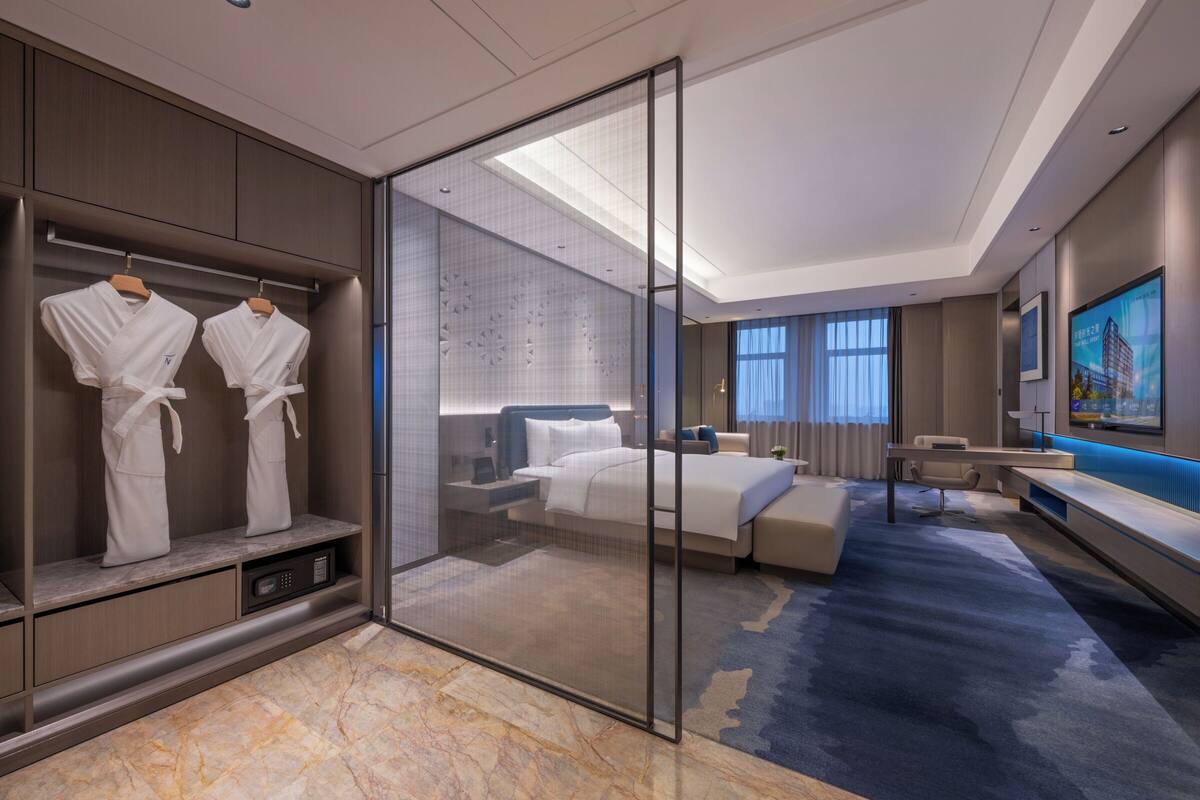A modern hotel room with a bed, a desk, a seating area, and a walk-in closet with robes.