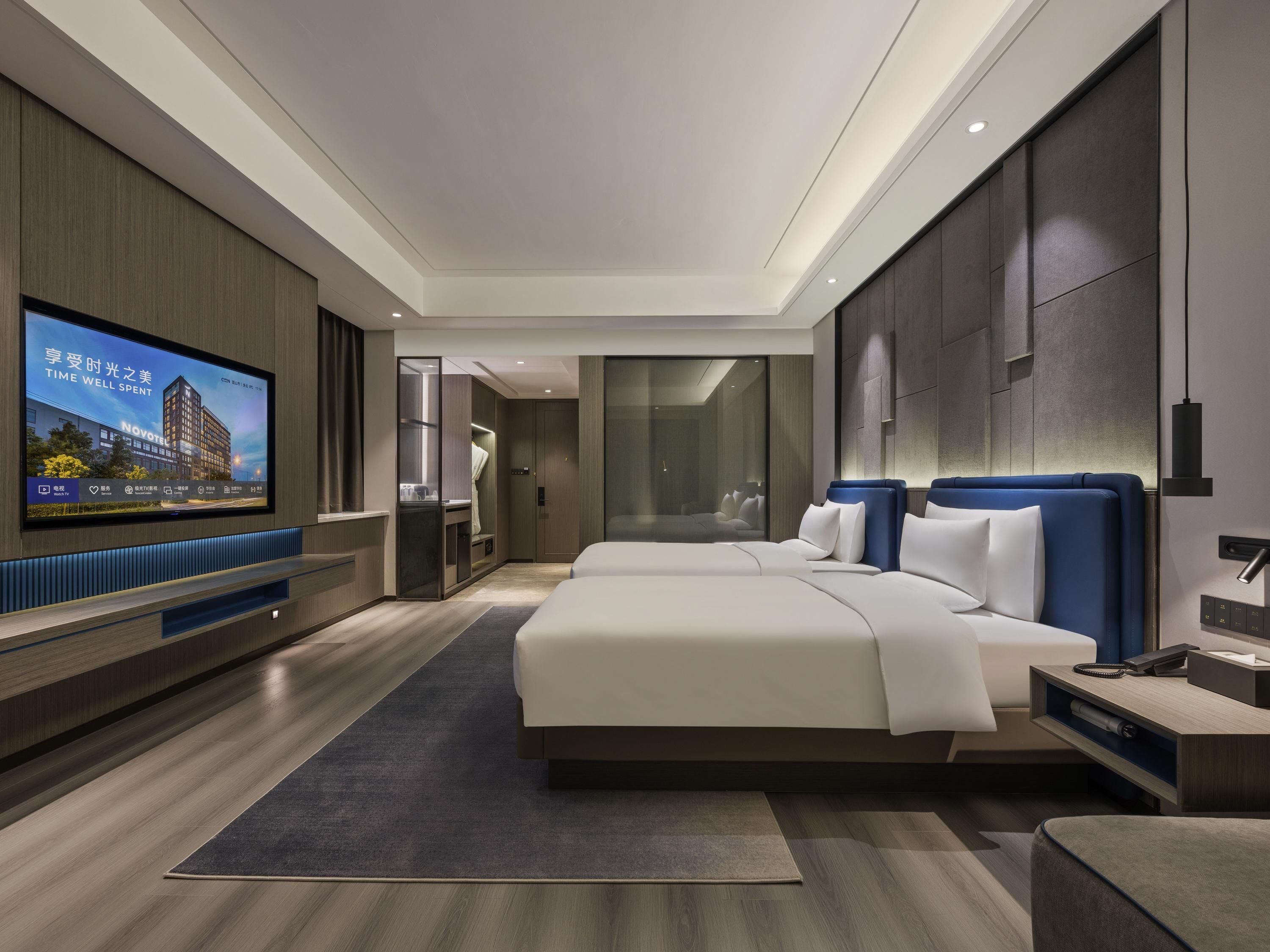 Een moderne hotelkamer met een grote flatscreen televisie, een bed met blauwe hoofdborden, een nachtkastje en een inbouwkleerkast.