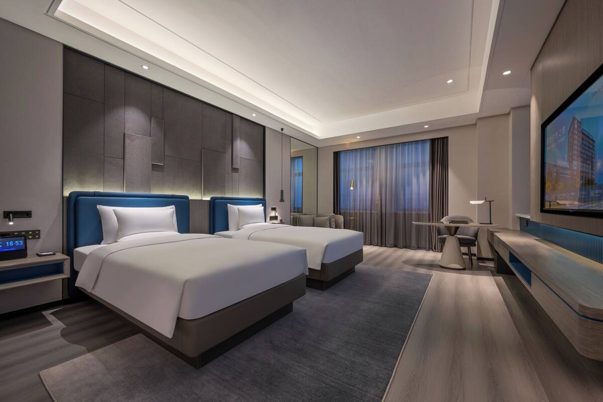 A modern hotel room with two beds, a desk, a chair, a TV, and a large window with curtains.