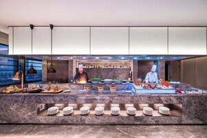 A modern kitchen with chefs preparing food, marble countertops, and a variety of dishes displayed.