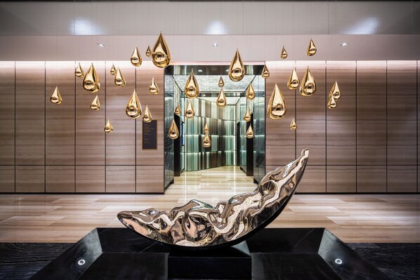 Een moderne lobby met een reflecterend sculptuur, een spiegelwand en een strakke liftruimte.