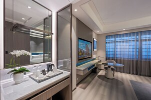 Quarto de hotel moderno com um espelho grande, televisão de tela plana, escrivaninha com cadeira, planta em vaso e uma janela ampla com cortinas.