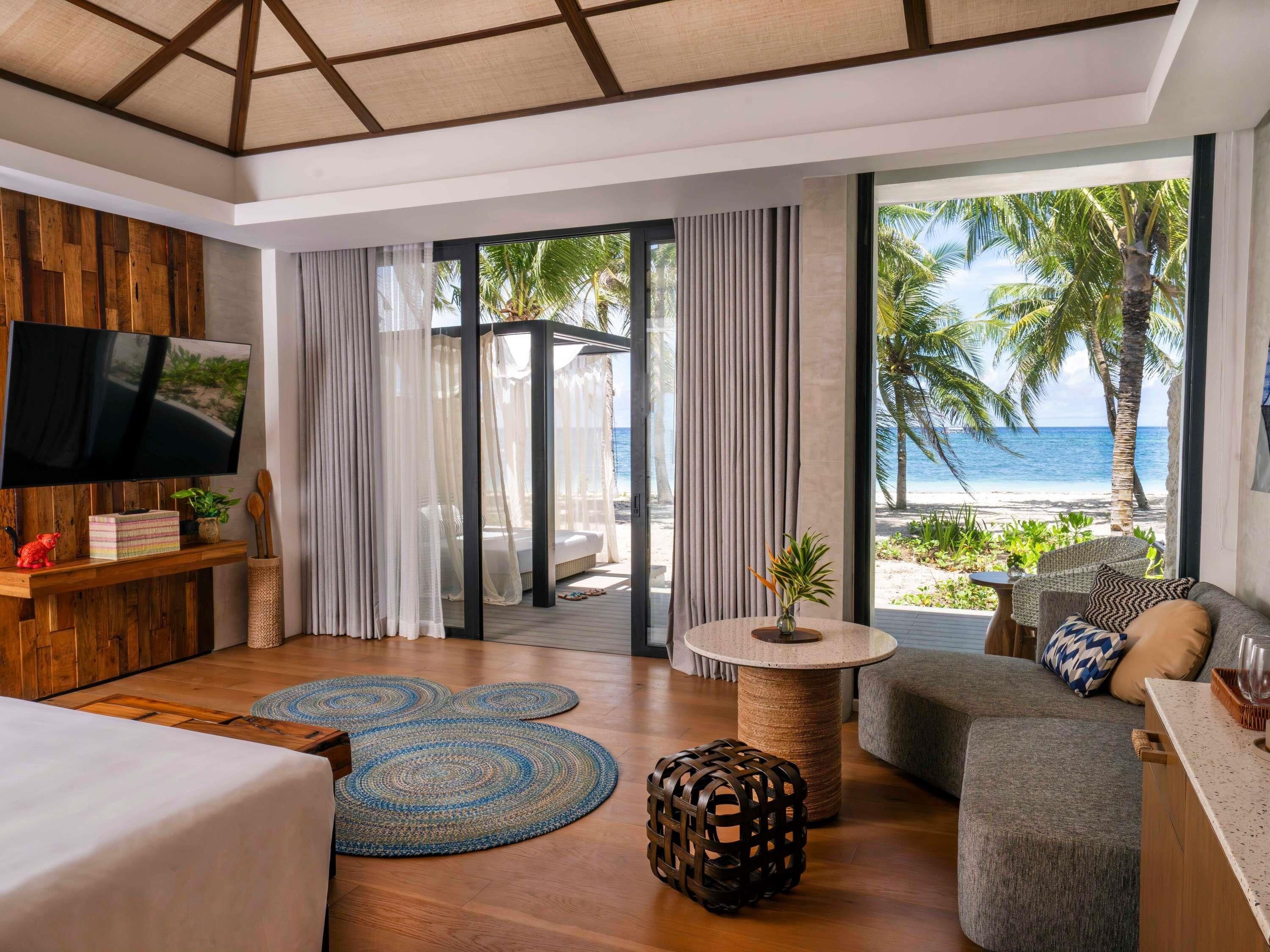 Una habitación con cama, sofá, televisor, una pequeña mesa con un florero y vista a la playa.