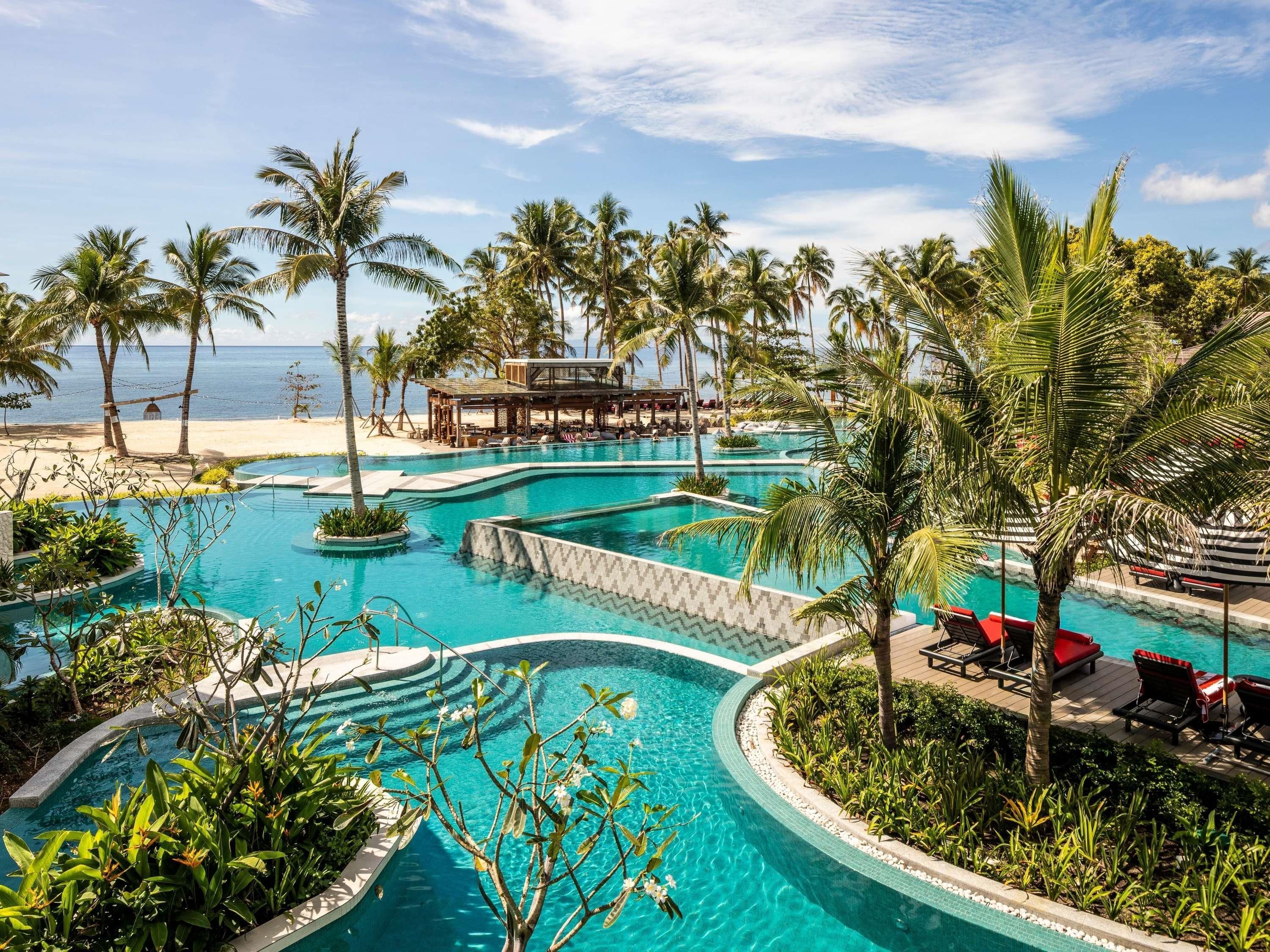 A resort pool with multiple sections, surrounded by palm trees and a beach view.