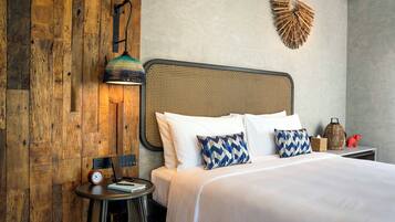 Habitación de hotel con pared de madera, cama con almohadillas de colores blanco y azul, cabecero tejido, una mesita con un reloj y un libro, y un adorno en la pared.