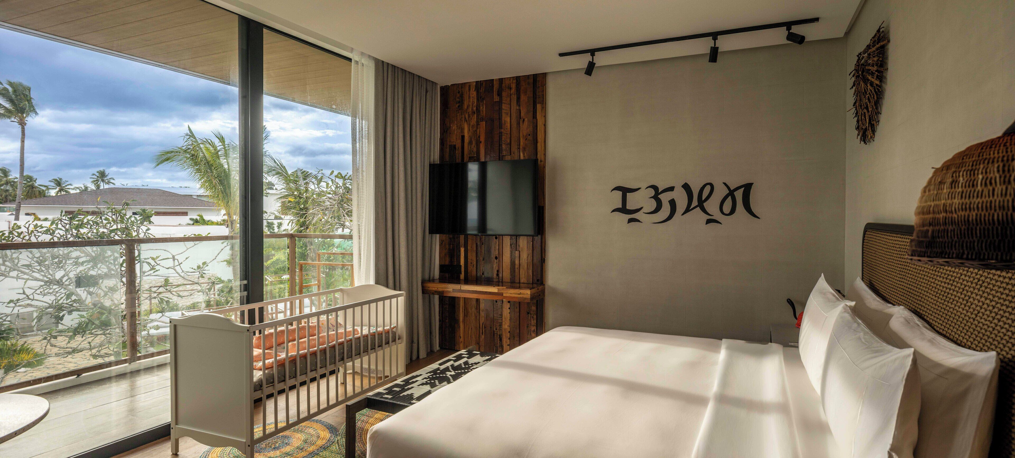 A hotel room with a large bed, a TV mounted on the wall, a crib, and a balcony with a view of palm trees and a building.