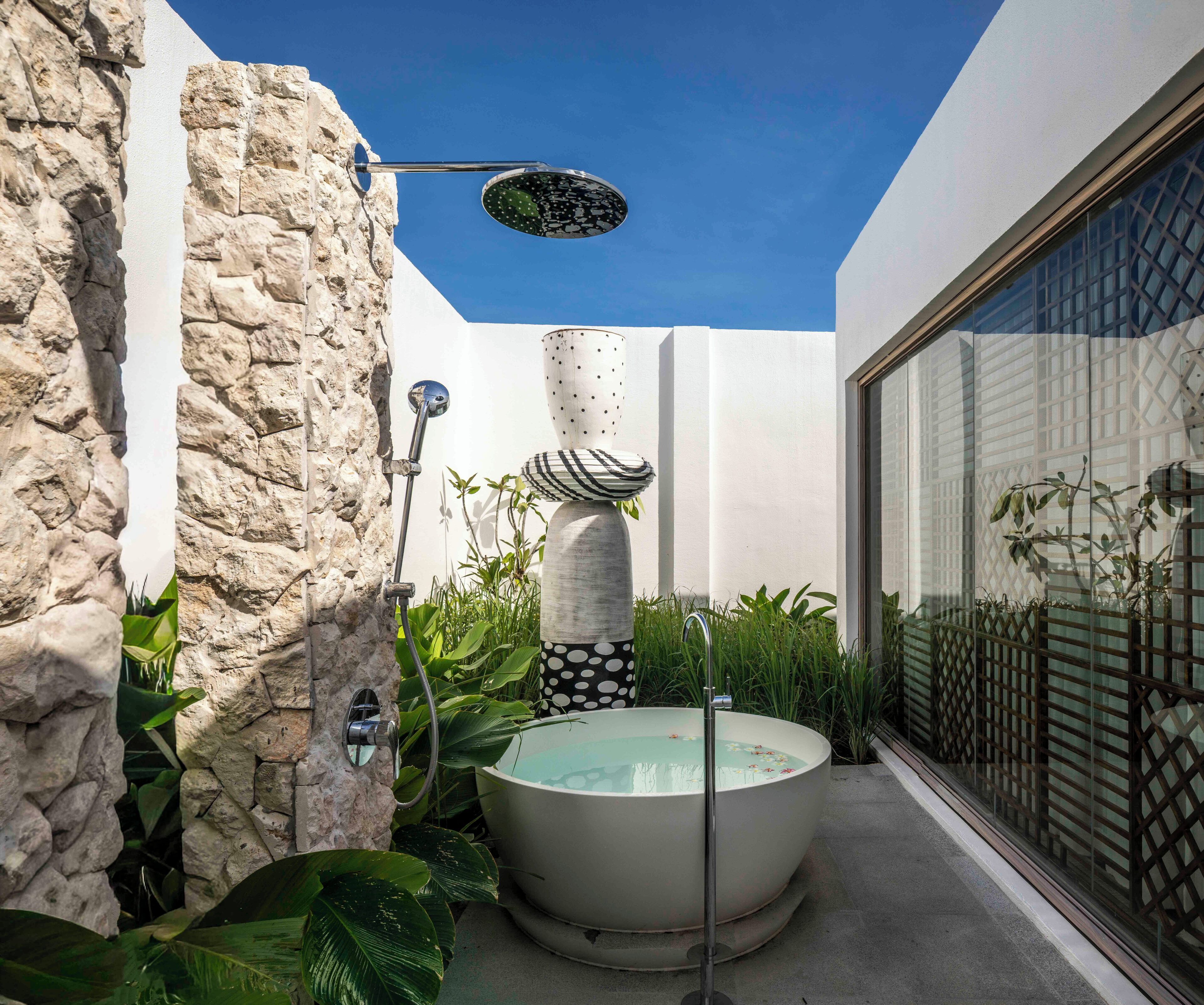 Un baño al aire libre moderno con una bañera independiente, ducha de lluvia y una pared de piedra.