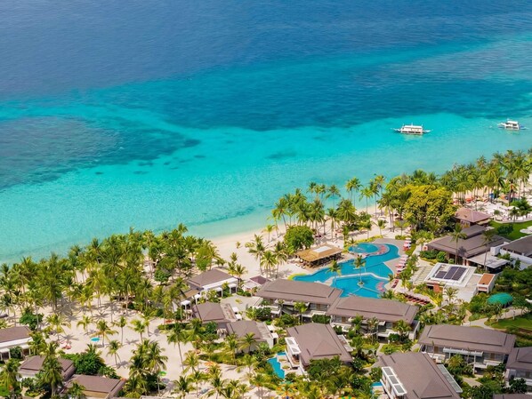 Un complexe hôtelier comprenant plusieurs bâtiments, une piscine et une plage bordée d’une eau turquoise et cristalline.