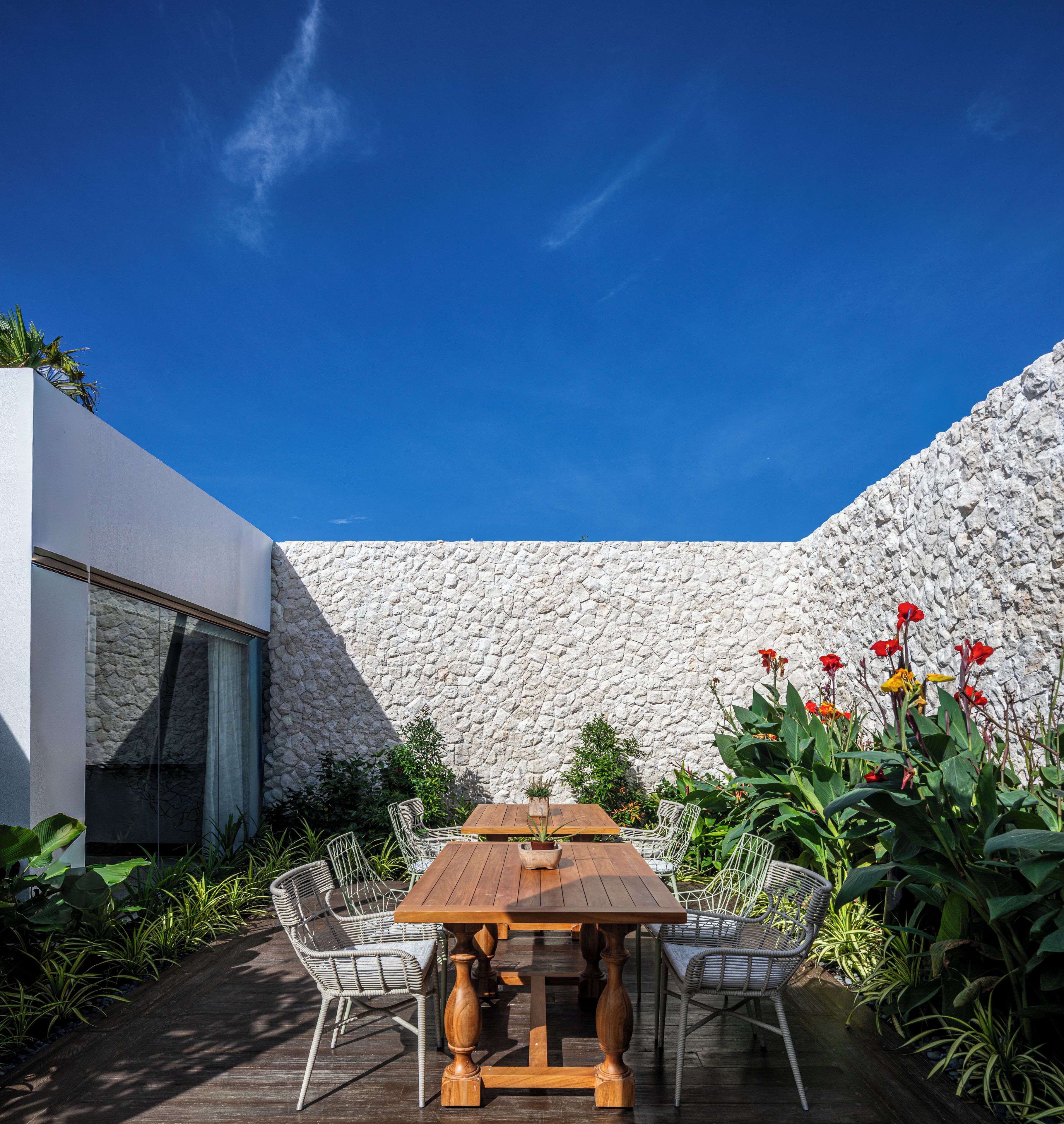 An outdoor dining area with a stone wall, wooden table, wicker chairs, and potted plants.