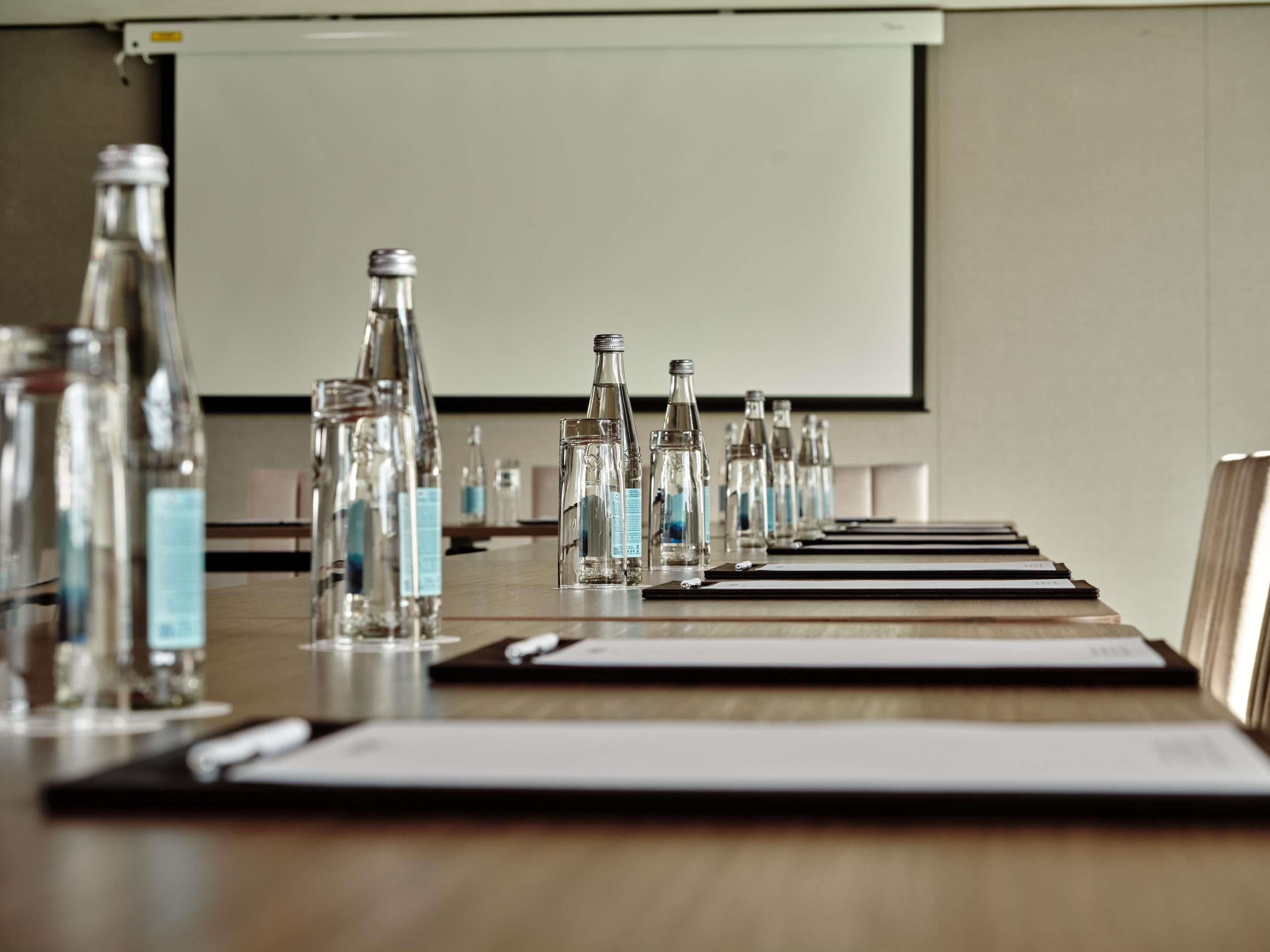 A conference table with water bottles and nameplates, a projector screen, and a whiteboard in a meeting room.