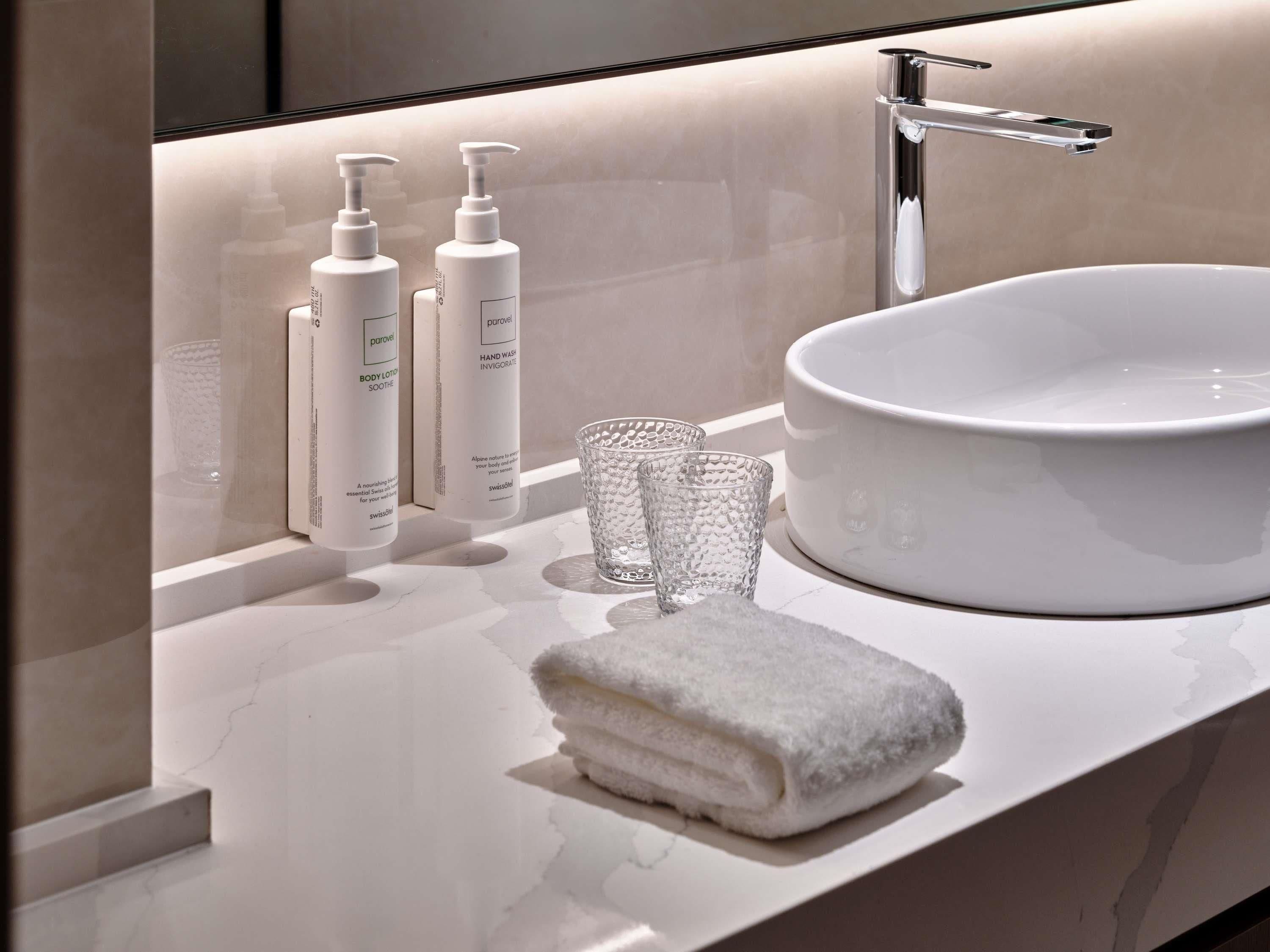 A modern bathroom with a white sink, two bottles of body lotion, a glass, and a folded towel on the counter.