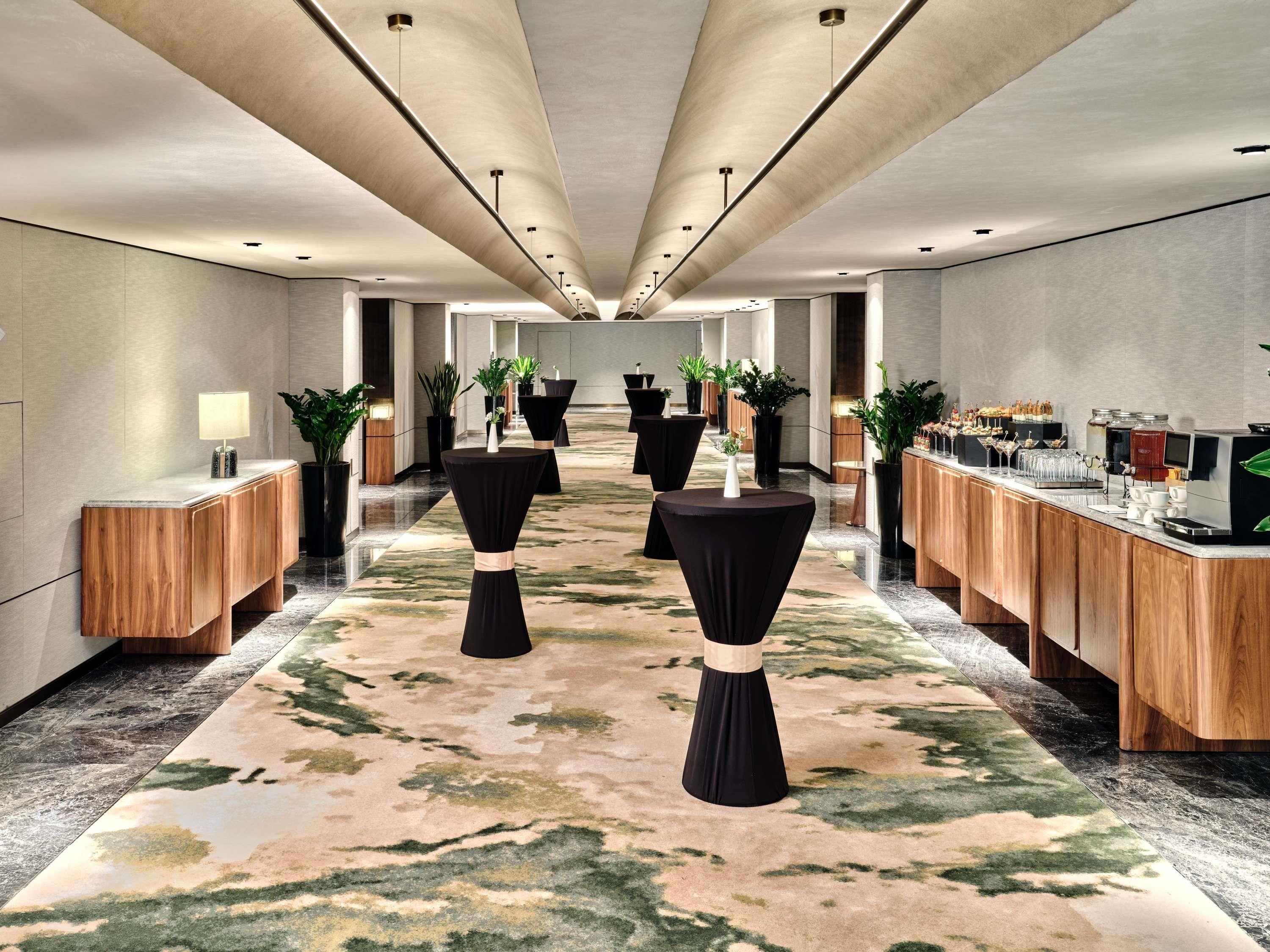 A hotel lobby with a central corridor, round tables, a coffee station, and potted plants.