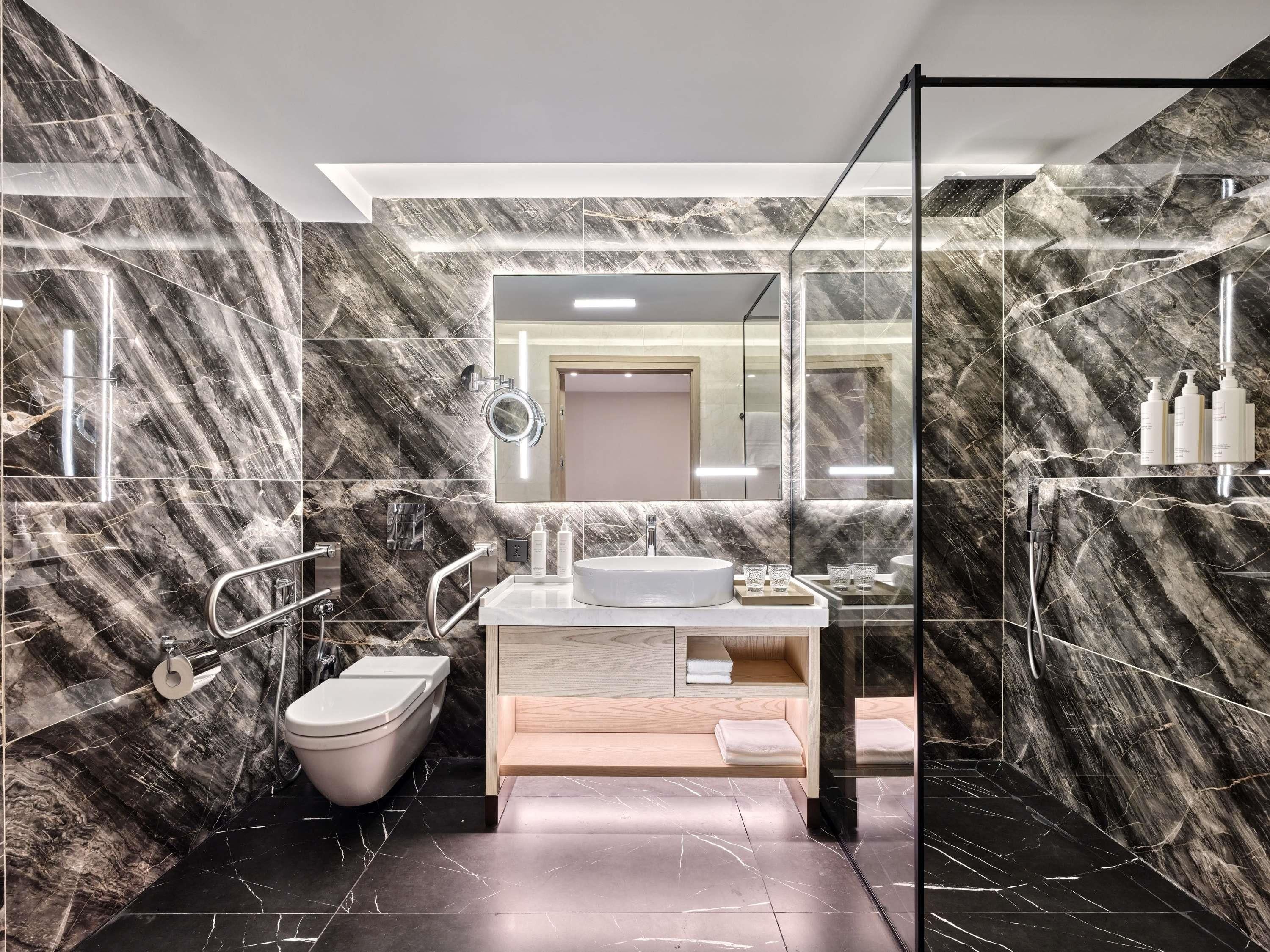 A modern bathroom with marble walls, a round mirror, a toilet, a sink, and a shower area.
