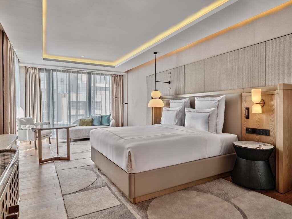 A modern hotel room with a large bed, bedside tables, a desk, and a view of the city.