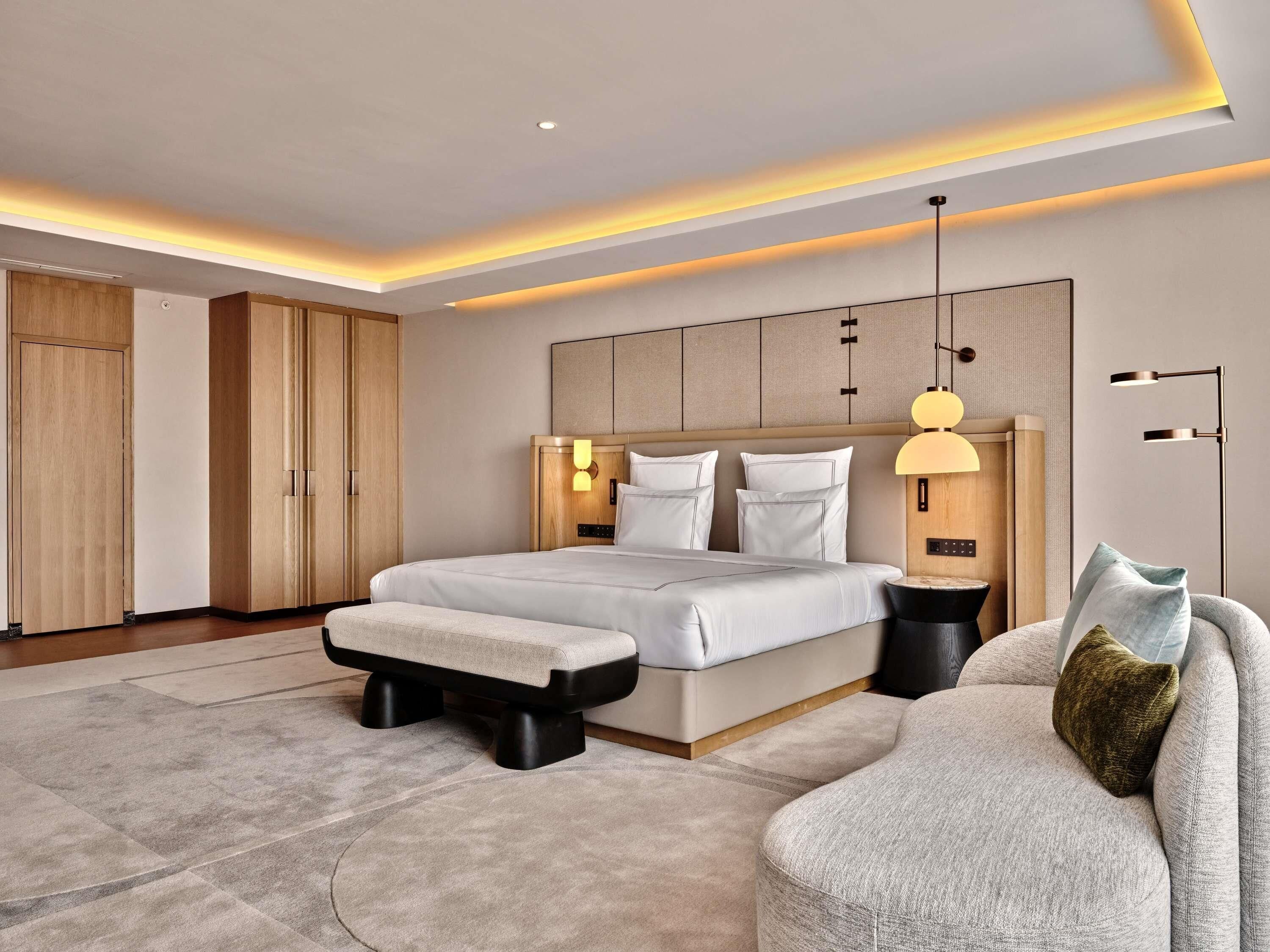 A modern bedroom with a large bed, bedside tables, a sofa, and a headboard with integrated lighting.