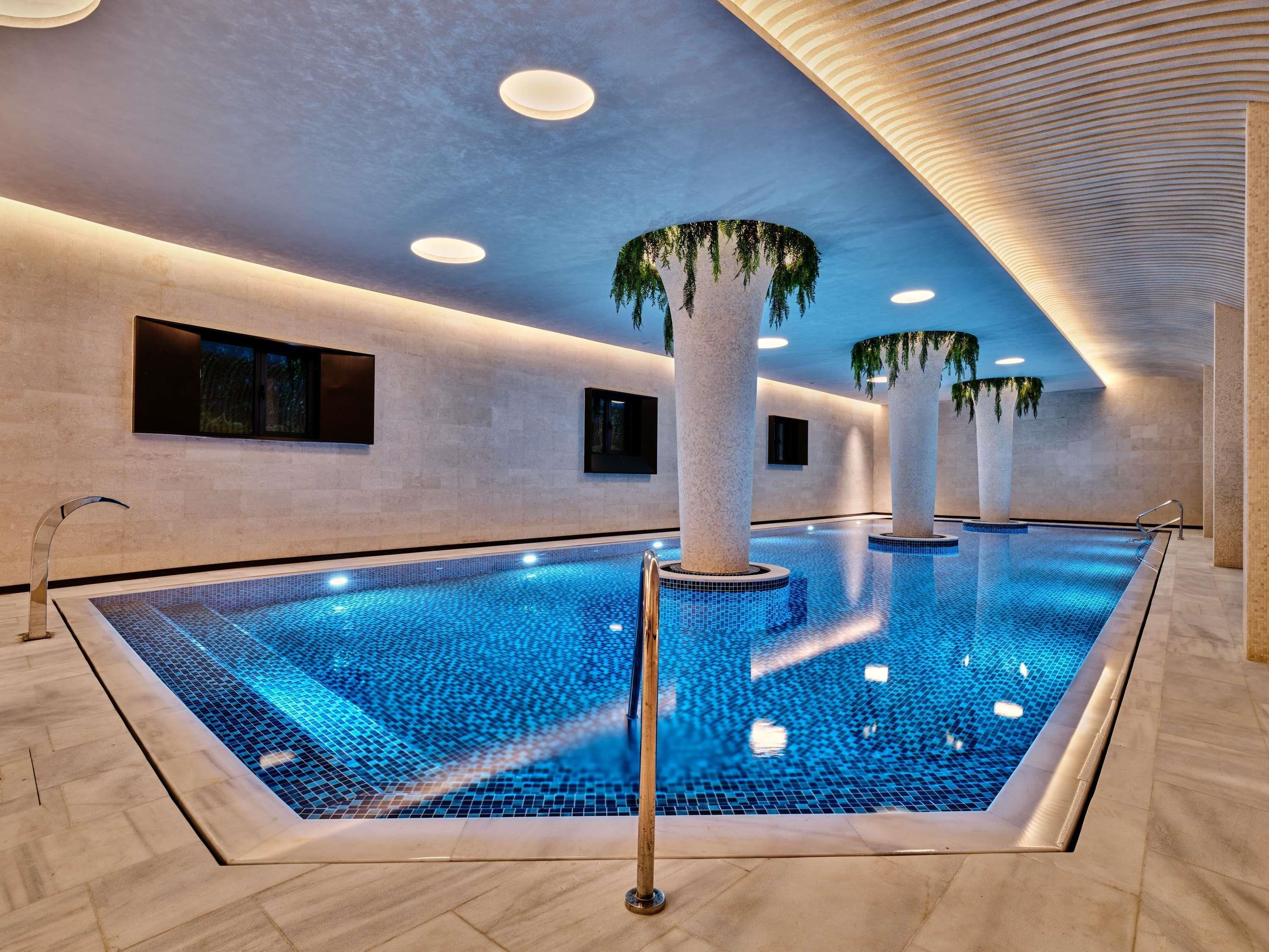 Une piscine intérieure dotée d’un sol carrelé, de colonnes ornées de plantes, d’un éclairage encastré et d’un design de plafond moderne.