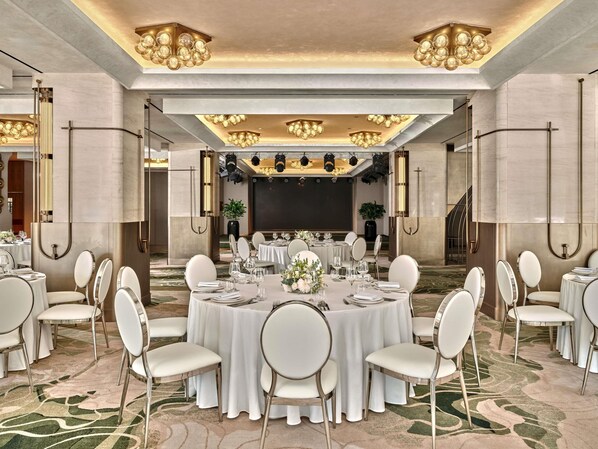A banquet hall with round tables, white chairs, and chandeliers.