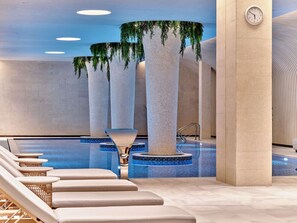 Un área de piscina interior moderna con un elemento acuático central, sillas de descanso y un reloj en la pared.