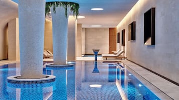 Una piscina interior moderna con baldosas azules cristalinas, amplias columnas y sillones de descanso a lo largo de las paredes.