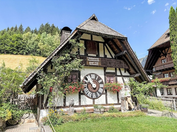 A traditional half-timbered building with a large clock, surrounded by greenery and a clear sky.