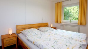 Un dormitorio con una cama de madera, dos mesitas de noche, una lámpara de noche, una ventana con cortinas y un radiador.