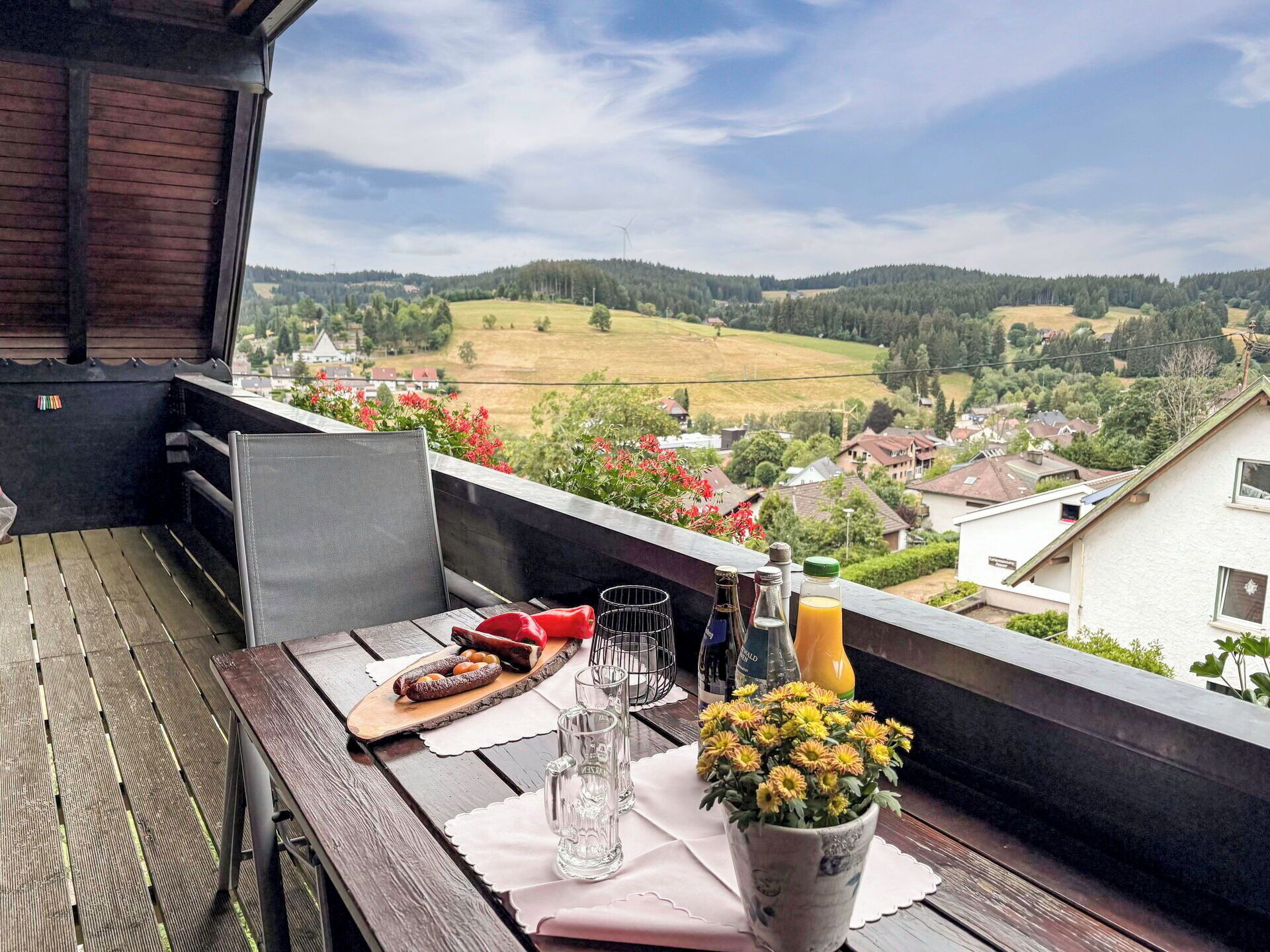 A balcony with a wooden table set for dining, a bottle of wine, glasses, and a flower vase.