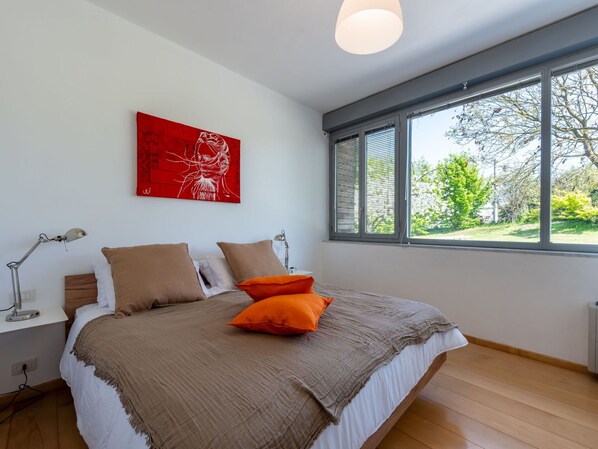 A bedroom with a bed, two bedside lamps, a red abstract painting, and a window with a view of trees.