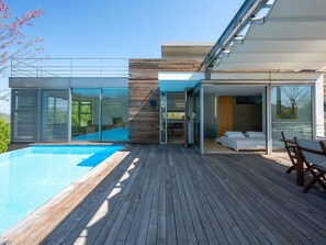 A modern house with a swimming pool, wooden deck, and glass doors leading to a bedroom.