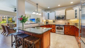 A modern kitchen with a large island, stainless steel appliances, and a dining area with wicker chairs. - Regency Villas 212 Lush Garden By Suite Paradise (Koloa)