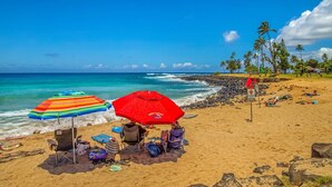 A beach with two colorful umbrellas, a chair, and a sign in the background. - Regency Villas 212 Lush Garden By Suite Paradise (Koloa)