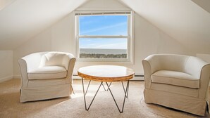 A room with a sloped ceiling, a large window, two white armchairs, and a round wooden table. - 2024 S. Bay Shore Dr. — Private, Peaceful & Perfect, Your Bayfront Broadkill Beach Escape Awaits! (Milton)