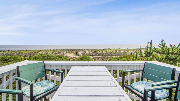 A deck with two green chairs and a white table, overlooking a beach and greenery. - 2024 S. Bay Shore Dr. — Private, Peaceful & Perfect, Your Bayfront Broadkill Beach Escape Awaits! (Milton)