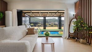 A modern living room with a sofa, a small table, and a view of a pool and mountains. - Yeongheungdo Stay Noc Pool Villa (Incheon)