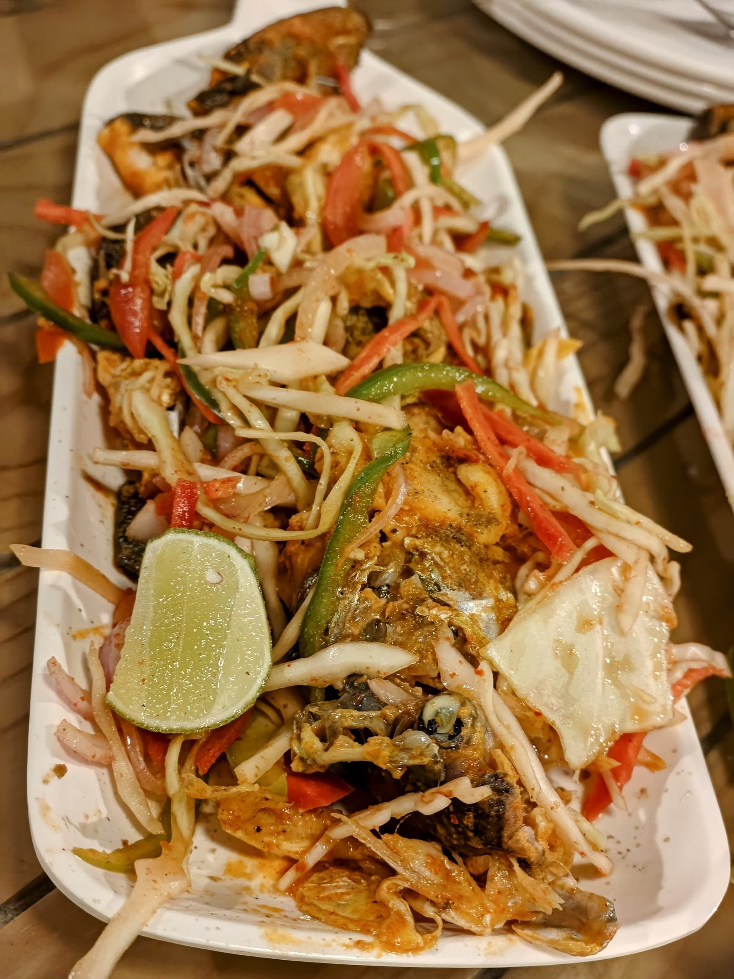A dish with fish, vegetables, and a lime wedge on a white plate.