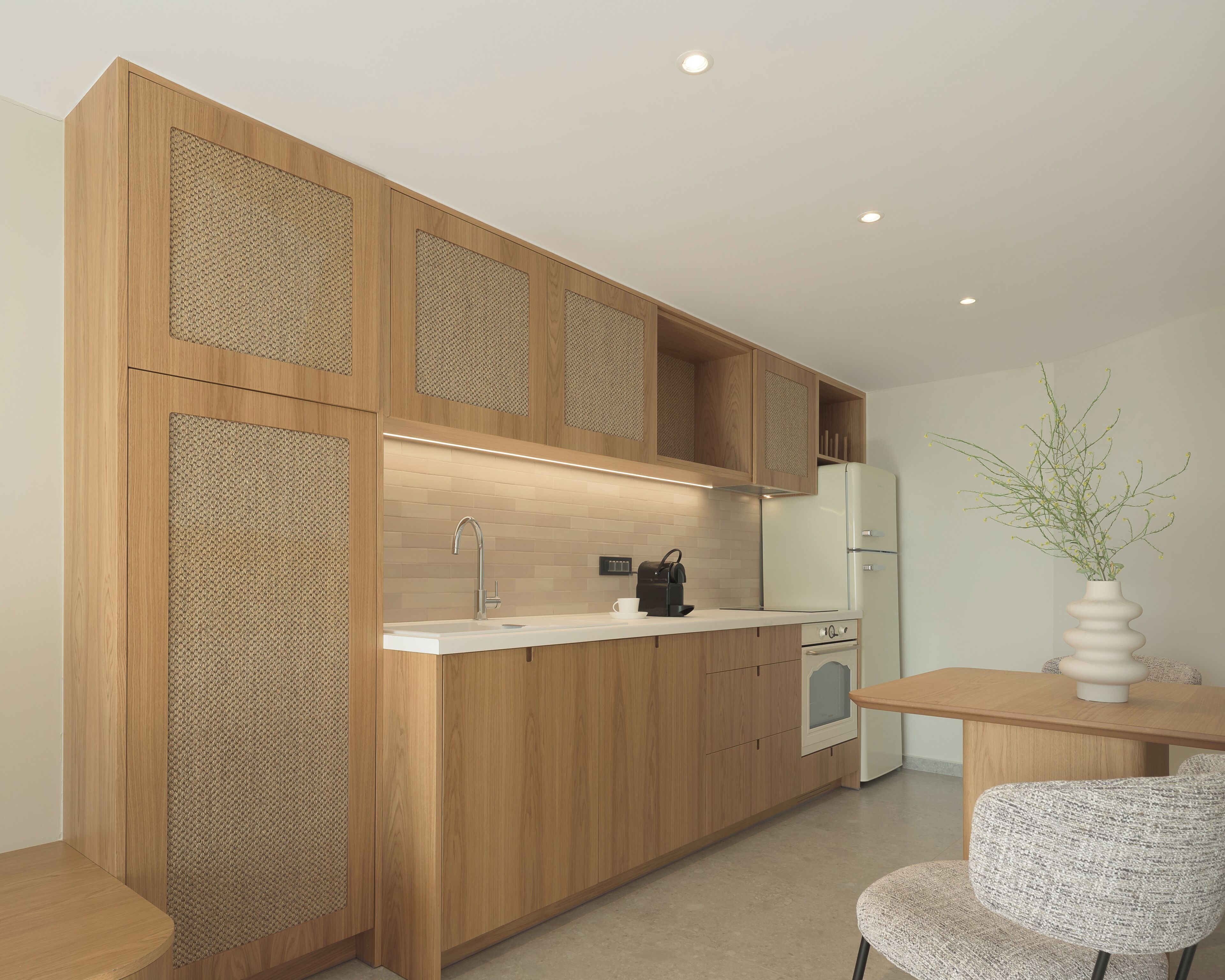 A modern kitchen with wooden cabinetry, a built-in sink, a coffee machine, and a dining area with a table and chair.