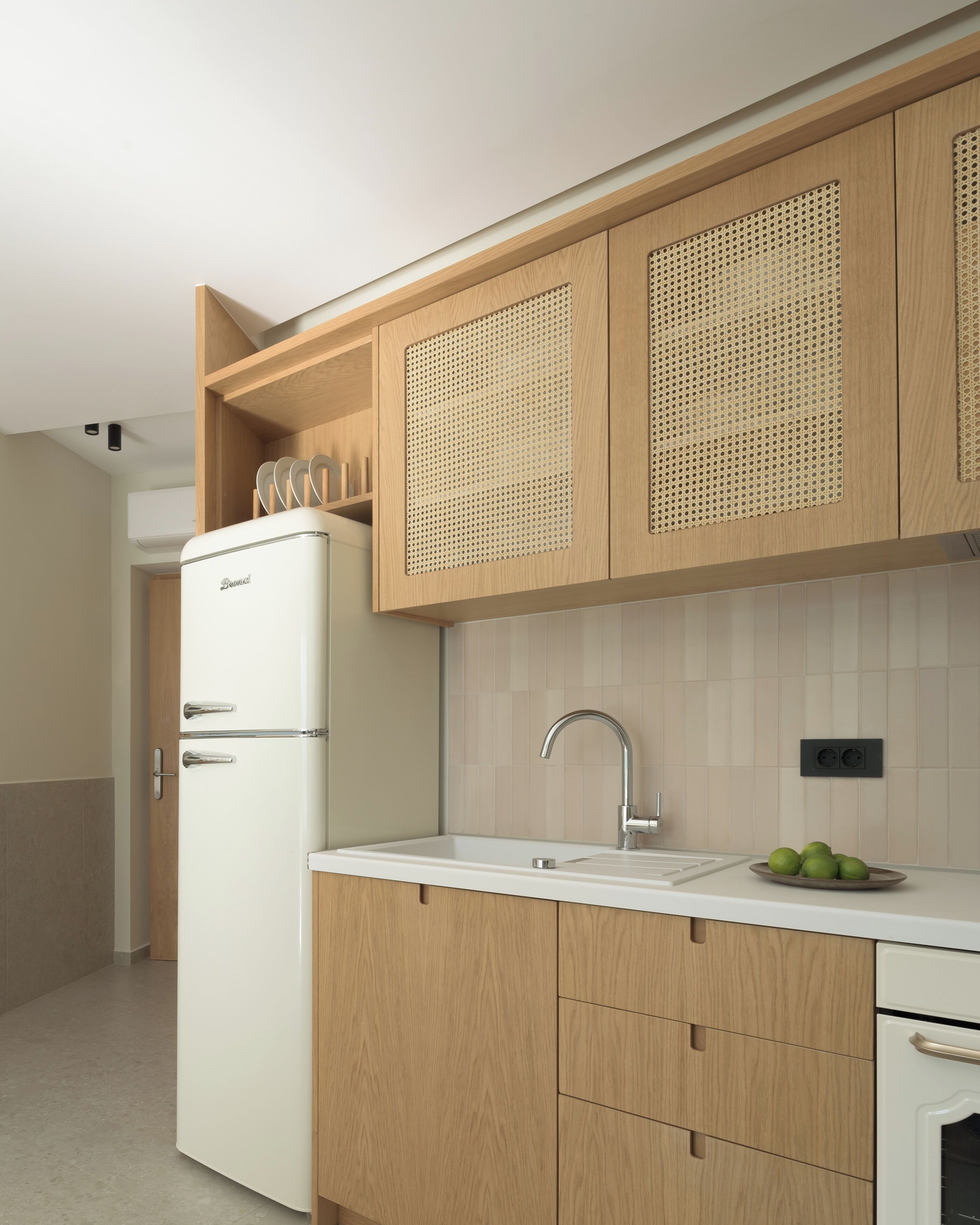 Une cuisine moderne avec des meubles en bois, un réfrigérateur blanc, un évier avec un robinet et un plan de travail sur lequel se trouve un saladier rempli de fruits.