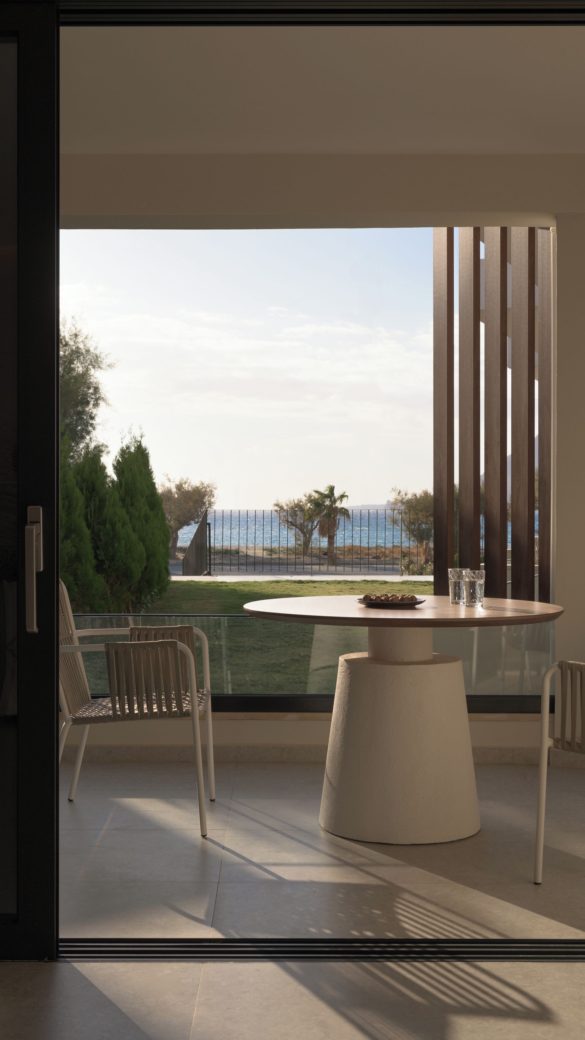 A modern balcony with a round table and chairs, overlooking a coastal landscape with a fence and trees.