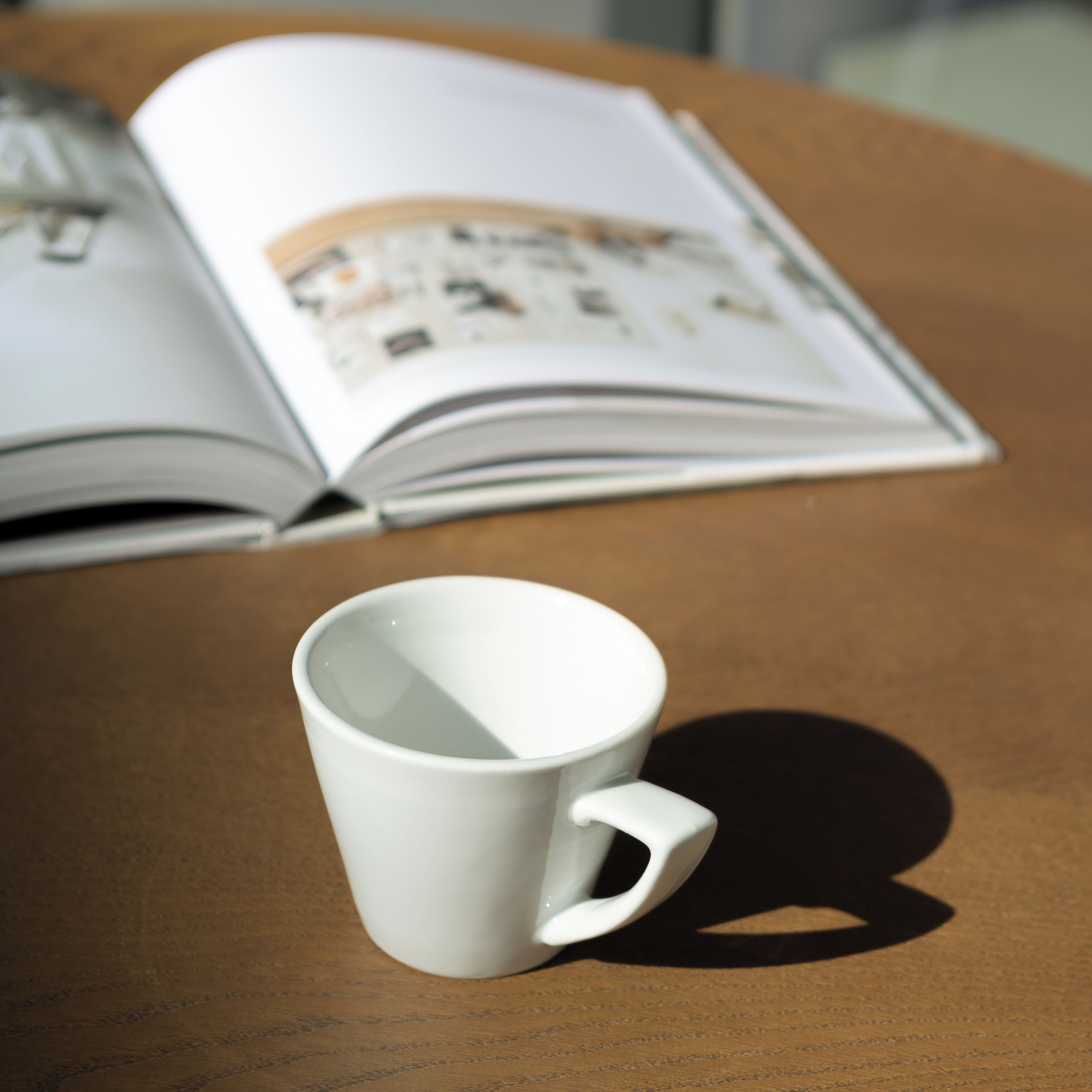 A white mug on a wooden table with an open book behind it.