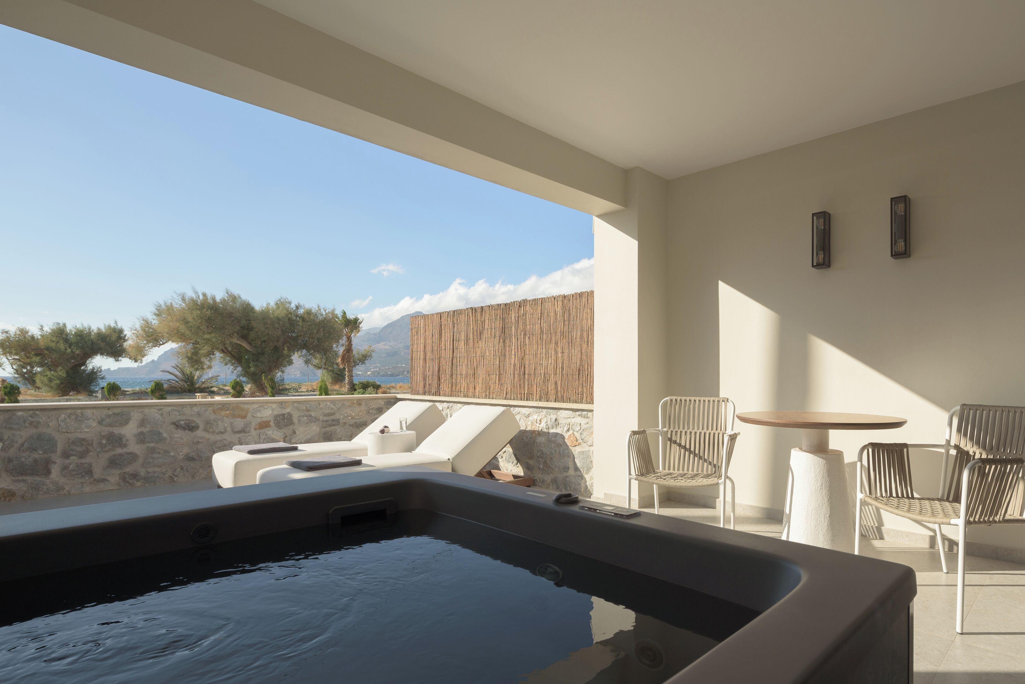 A modern outdoor hot tub area with lounge chairs and a table, overlooking a scenic landscape with mountains and trees.