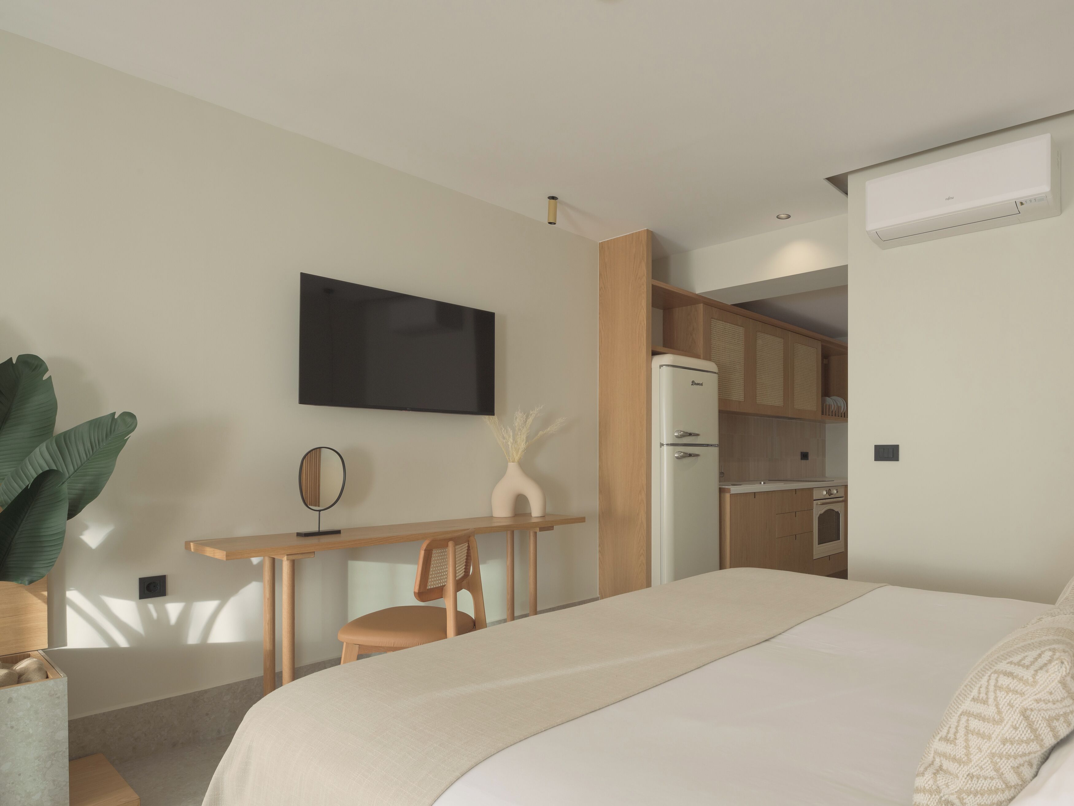 A modern hotel room with a bed, a desk, a chair, a flat-screen TV, a refrigerator, and a kitchenette.