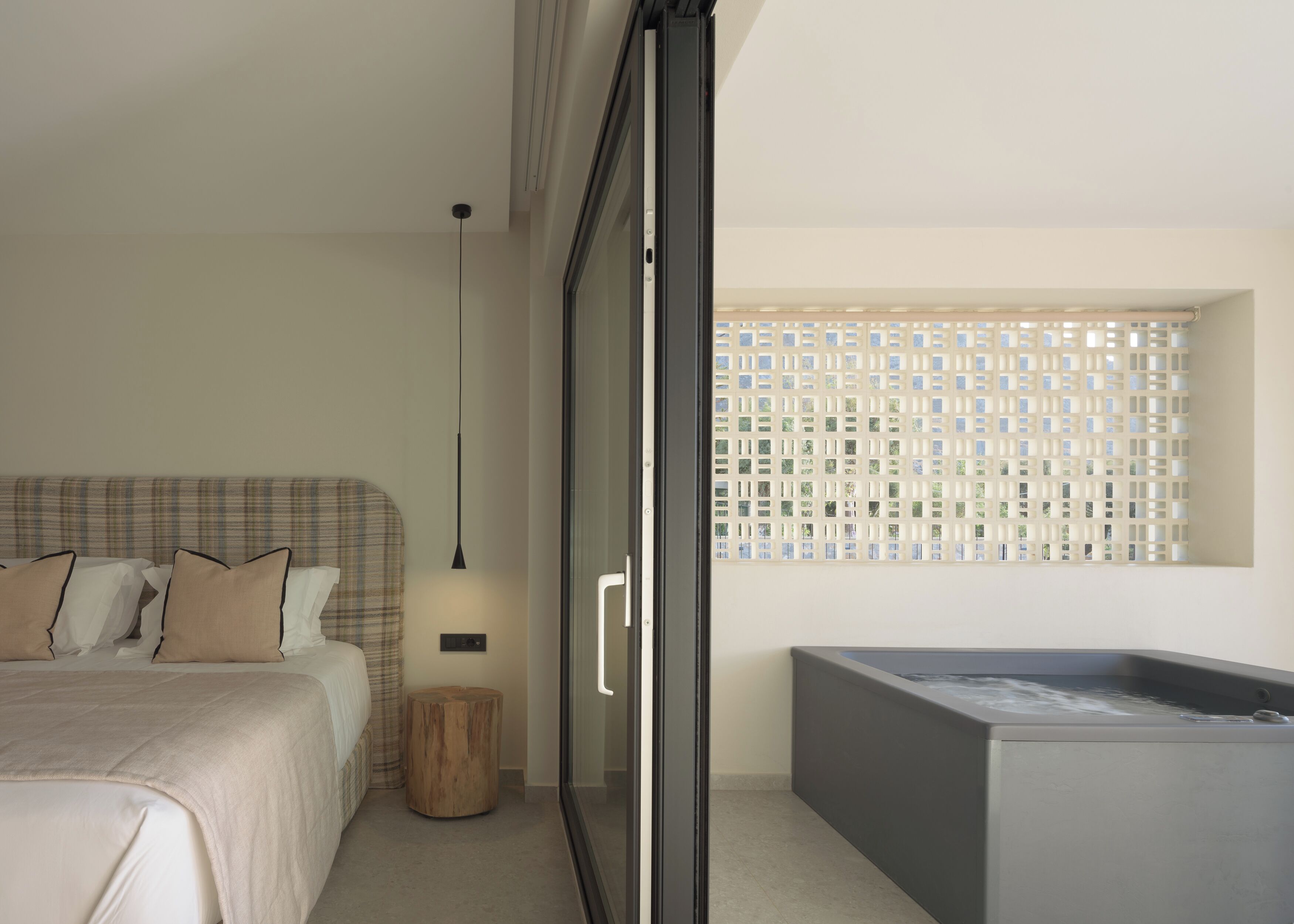 Un dormitorio moderno con una cama, un taburete de madera, una lámpara colgante negra y una bañera grande.