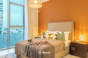 A modern bedroom with a large bed, a bedside table, a nightstand, a window with a view, and a hanging light fixture. - Manzil-1BR Beach & Lagoon Access (Dubai)