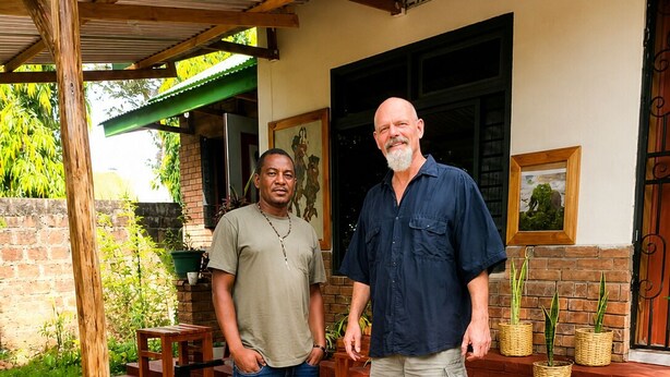 Two men standing on a porch with a brick wall, wooden furniture, and potted plants. - Kilimanjaro sons Homestay (Moshi)
