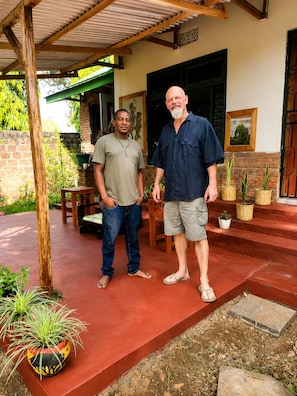 Two men standing on a porch with a brick wall, wooden furniture, and potted plants. - Kilimanjaro sons Homestay (Moshi)