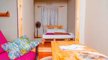 Een kamer met een bed, een ventilator, een tafel met een krant en een kleurrijk kussen.