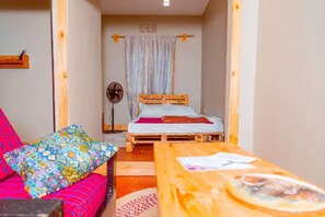 A room with a bed, a fan, a table with a newspaper, and a colorful cushion. - Kilimanjaro sons Homestay (Moshi)