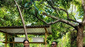 Two people standing in a garden with a wooden structure and leaf-covered ground. - Kilimanjaro sons Homestay (Moshi)